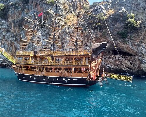 A large, ornate pirate-style ship named 'Grand Troys 07' floating in the bright turquoise waters near rocky cliffs, offering a scenic boat tour experience in Alanya.